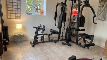 A home gym with a multi-functional exercise machine, a standing bike, and a small table with a lamp.