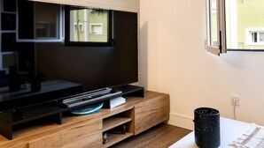 Ein modernes Wohnzimmer mit Flachbildfernseher, der an einer Holzwand angebracht ist, einem Fenster mit Jalousien und einem weißen Tisch mit dekorativem Teppich.