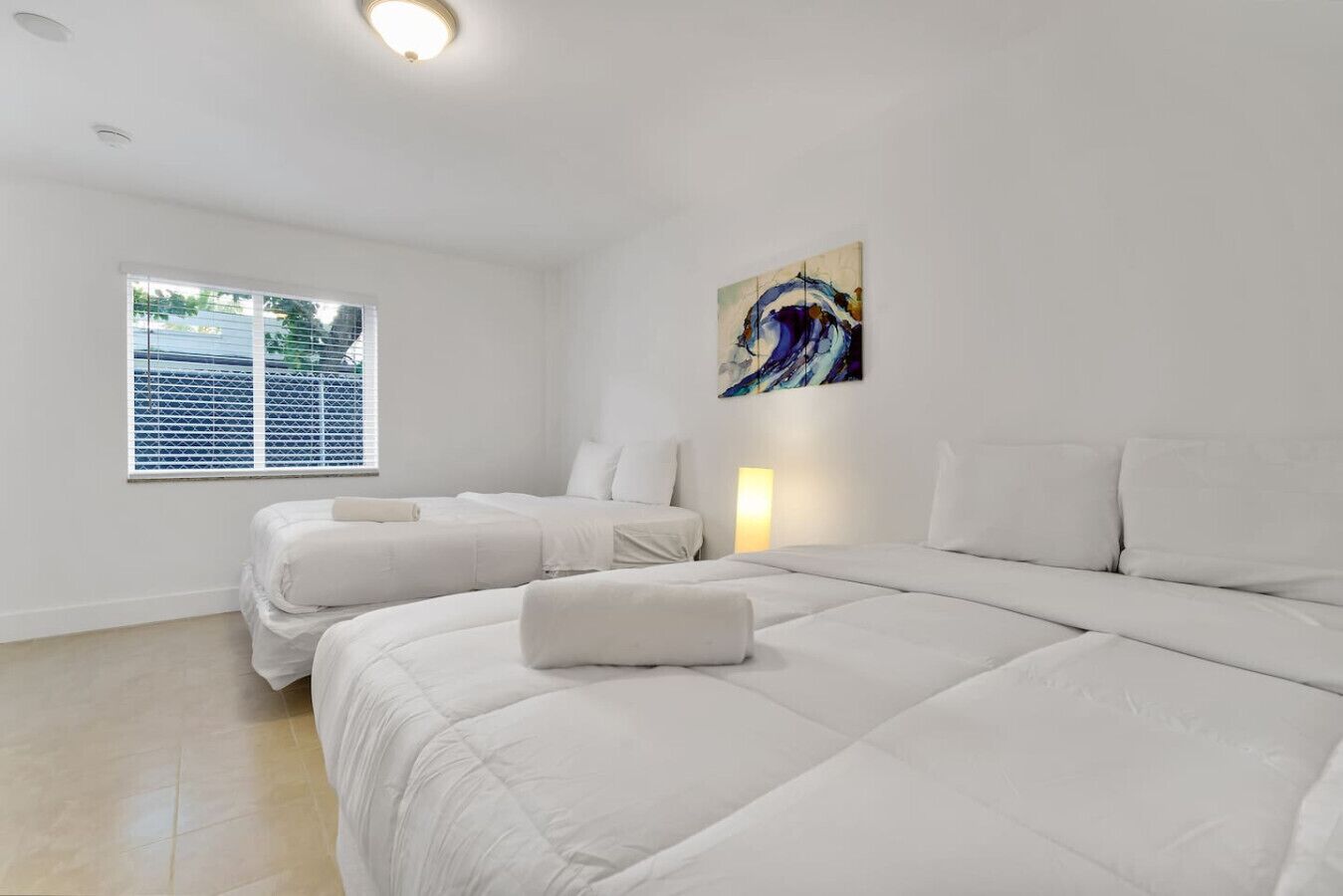 A hotel room with two beds, a window with blinds, a painting on the wall, and a bedside lamp.