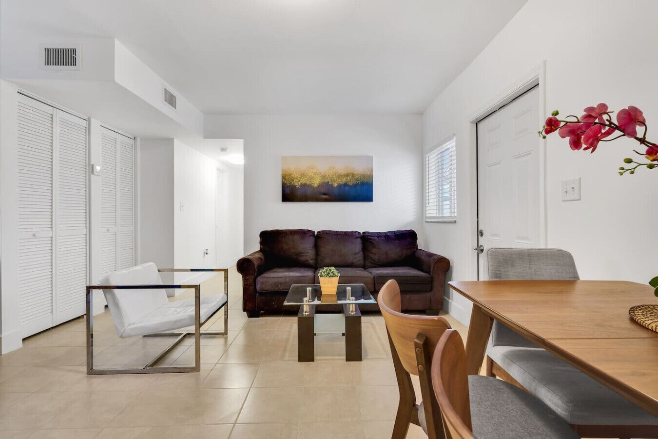 A modern living room with a sofa, coffee table, and dining table. There is a painting on the wall and a window with blinds.