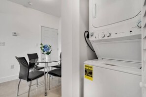 A modern hotel room with a dining area featuring a glass table and black chairs, a white door, and a white washing machine with an energy efficiency label. - ° Charming 2BR Apartment 5 min to Brickell ° (Miami)