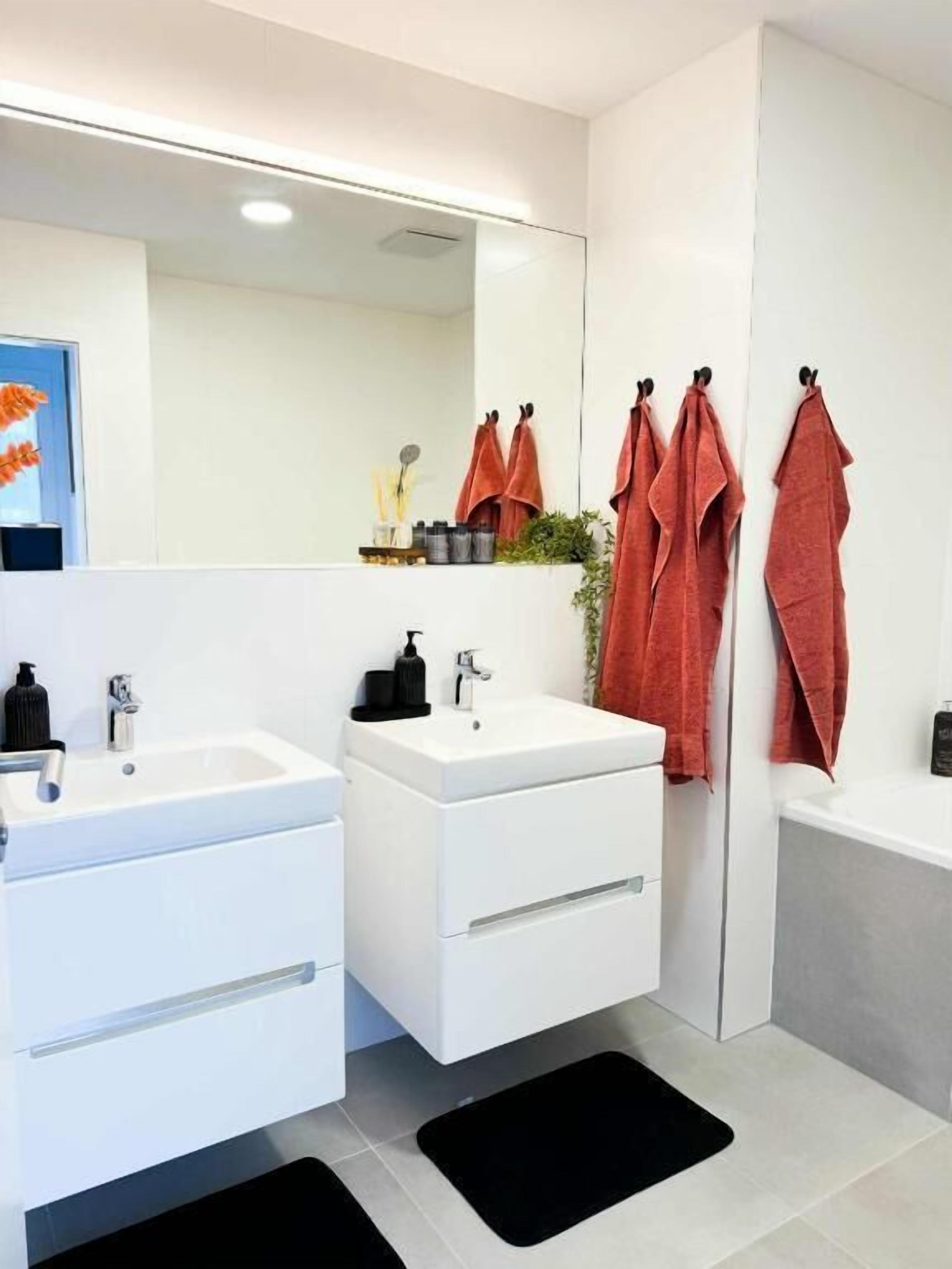 Ein modernes Badezimmer mit zwei Waschbecken, einem Spiegel und einer Badewanne. Handtücher hängen an Haken und ein Ablagebereich enthält Pflegeartikel.
