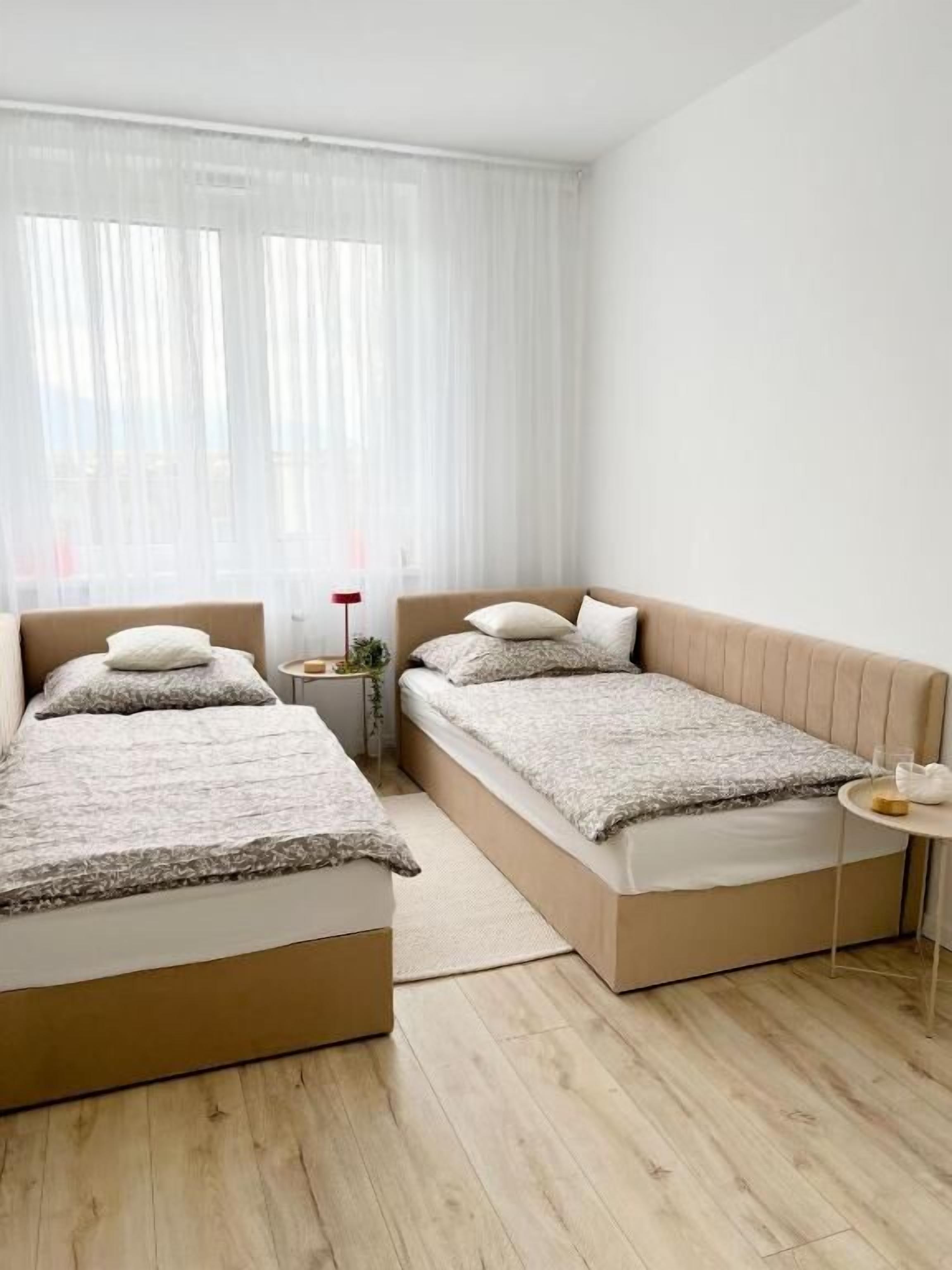A modern bedroom with two beds, a small bedside table, and a window with sheer curtains.
