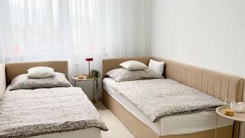 A modern bedroom with two beds, a small bedside table, and a window with sheer curtains.