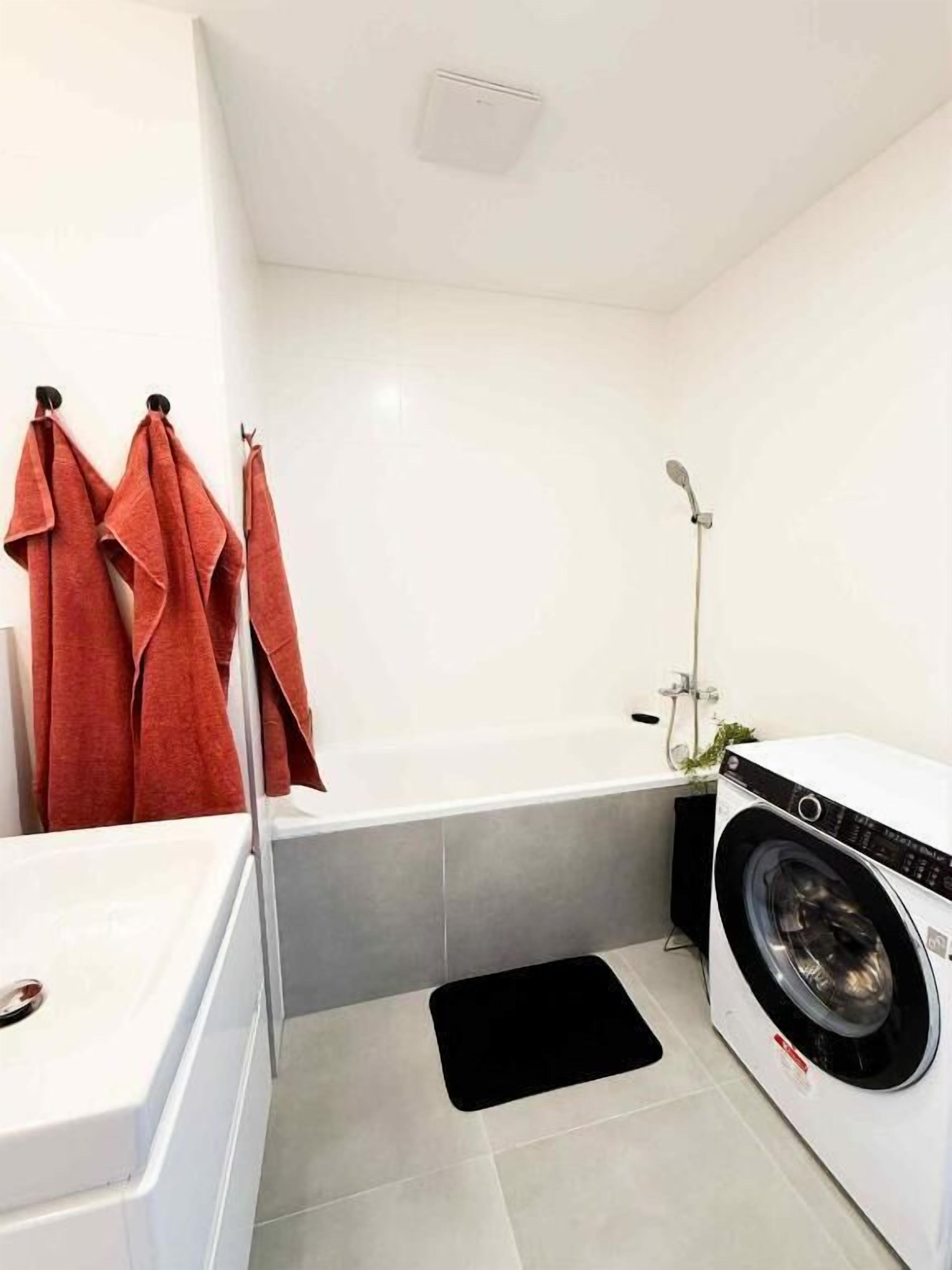 Een compacte badkamer met een wit bad, een wasmachine en een zwarte mat op de vloer.