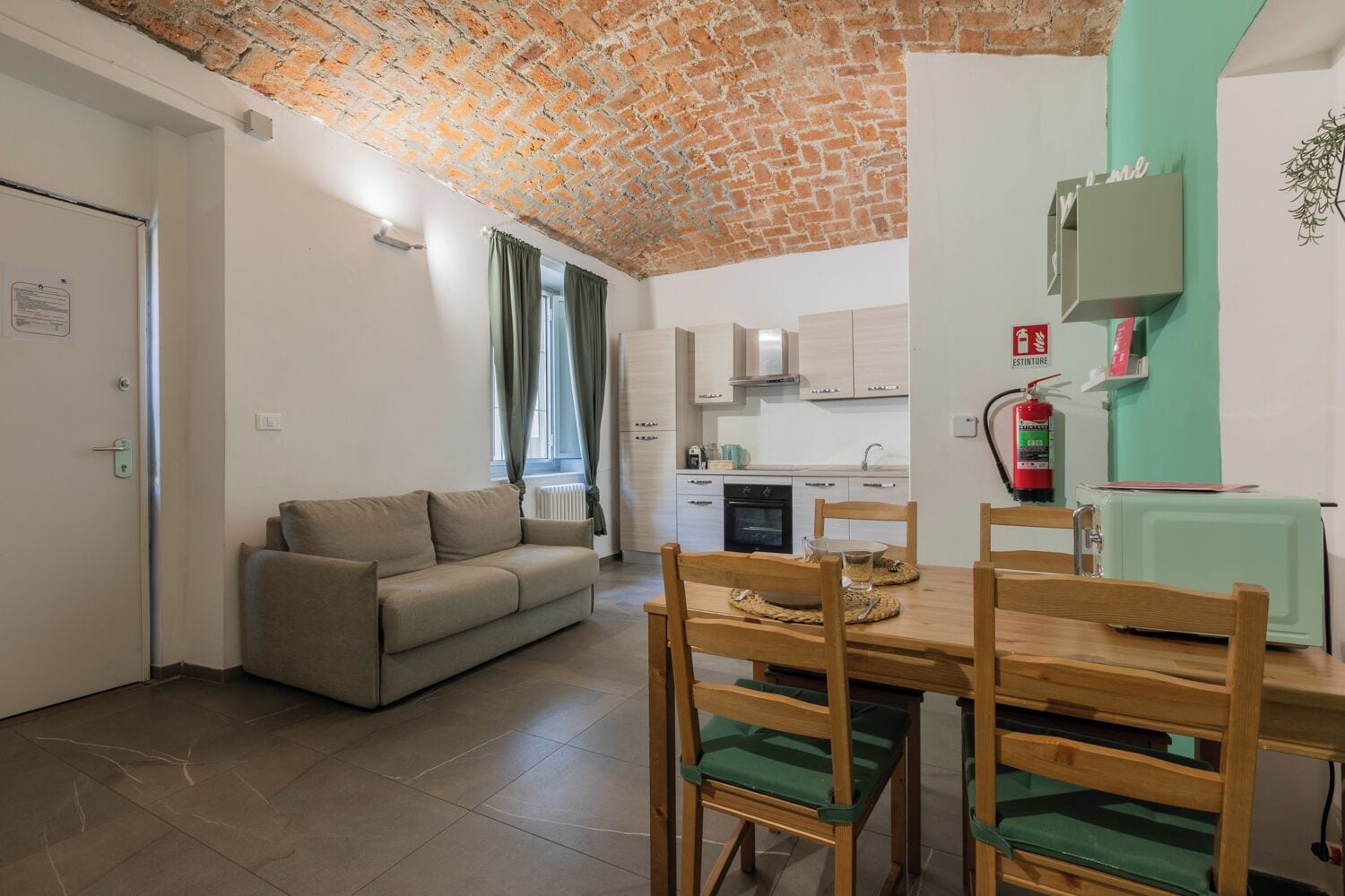 Ein Wohnzimmer mit Sofa, Esstisch und Stühlen, Küchenzeile mit Schränken, Ziegeldecke und Feuerlöscher.