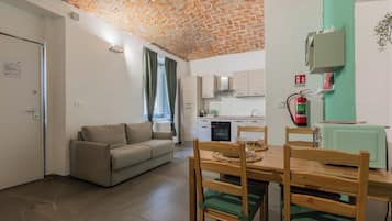 Ein Wohnzimmer mit Sofa, Esstisch und Stühlen, Küchenzeile mit Schränken, Ziegeldecke und Feuerlöscher.