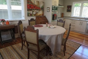 A dining area with a round table, chairs, a rug, a window with blinds, and a kitchen in the background. - Magpie Cottage - Homestay B&B (Taumarunui)