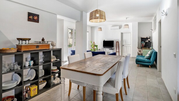 Un coin repas comprenant une table en bois, des chaises, un luminaire suspendu et une étagère où sont rangés de la vaisselle et des verres.