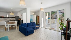 Un salon moderne avec un canapé bleu, une table à manger et une petite table ronde. On aperçoit un coin cuisine au fond, et une porte-fenêtre coulissante qui donne sur un espace extérieur.