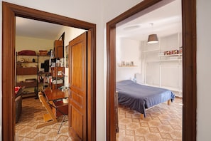 Ein Zimmer mit einem Bett, einem Schreibtisch und einem Stuhl. Eine Tür führt in einen weiteren Raum mit Regalen und einer Lampe.