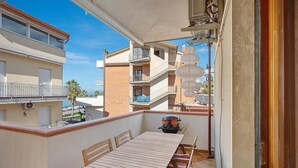 A balcony with a wooden table and chairs, a barbecue grill, and a view of neighboring buildings. - Levante Apartment with Sea View, Wi-Fi and Air Conditioning (Agropoli)