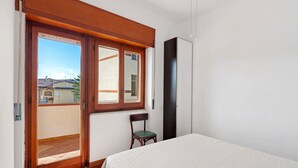 Ein Schlafzimmer mit einem Bett, einem Sessel, einem Kleiderschrank und einem Fenster mit Blick auf Gebäude.