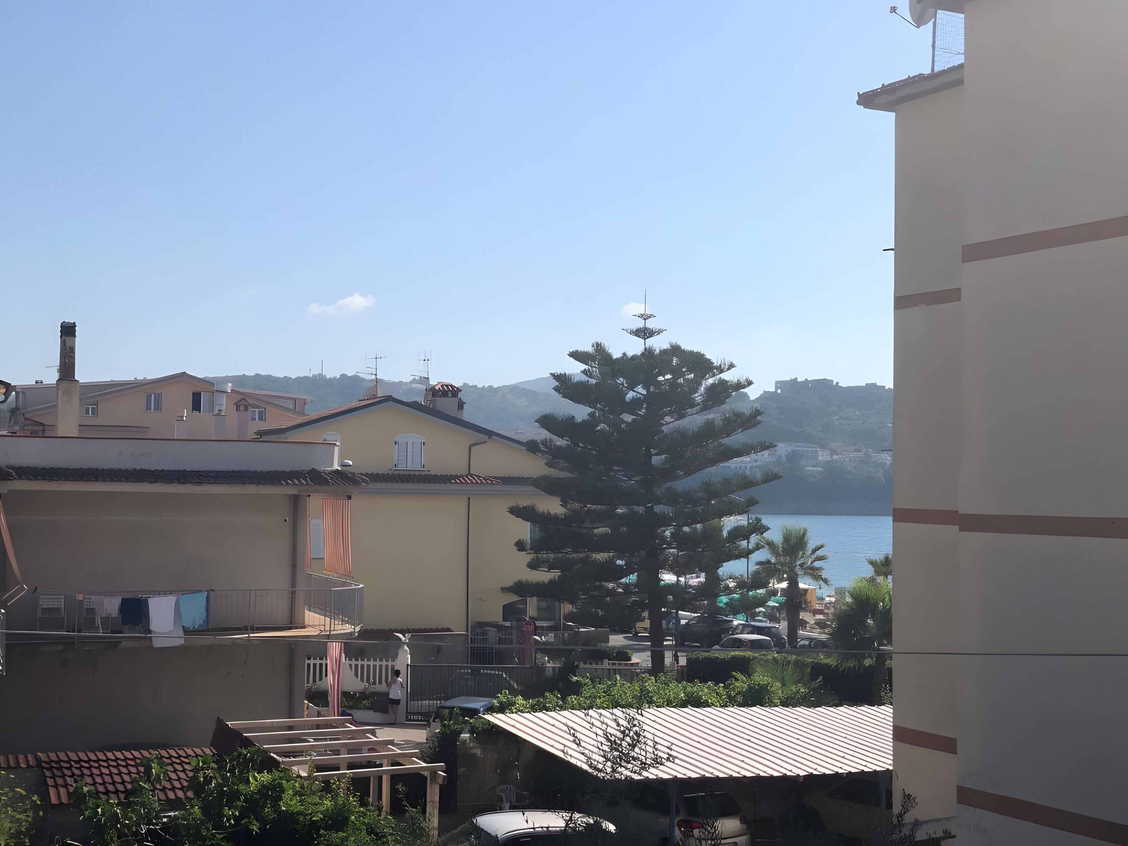 A residential area with a view of the sea, houses with tiled roofs, and a large tree.