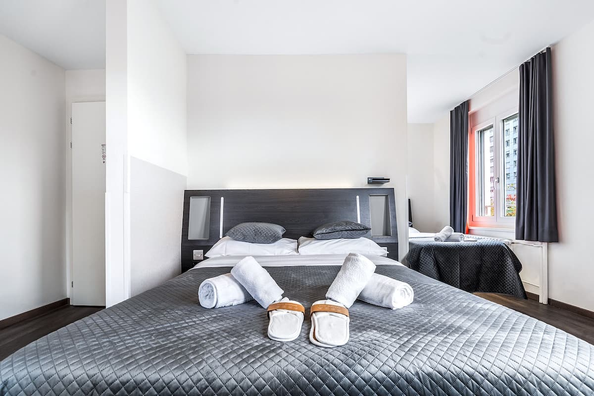A neatly made bed with a quilted grey cover, pillows, and rolled towels.