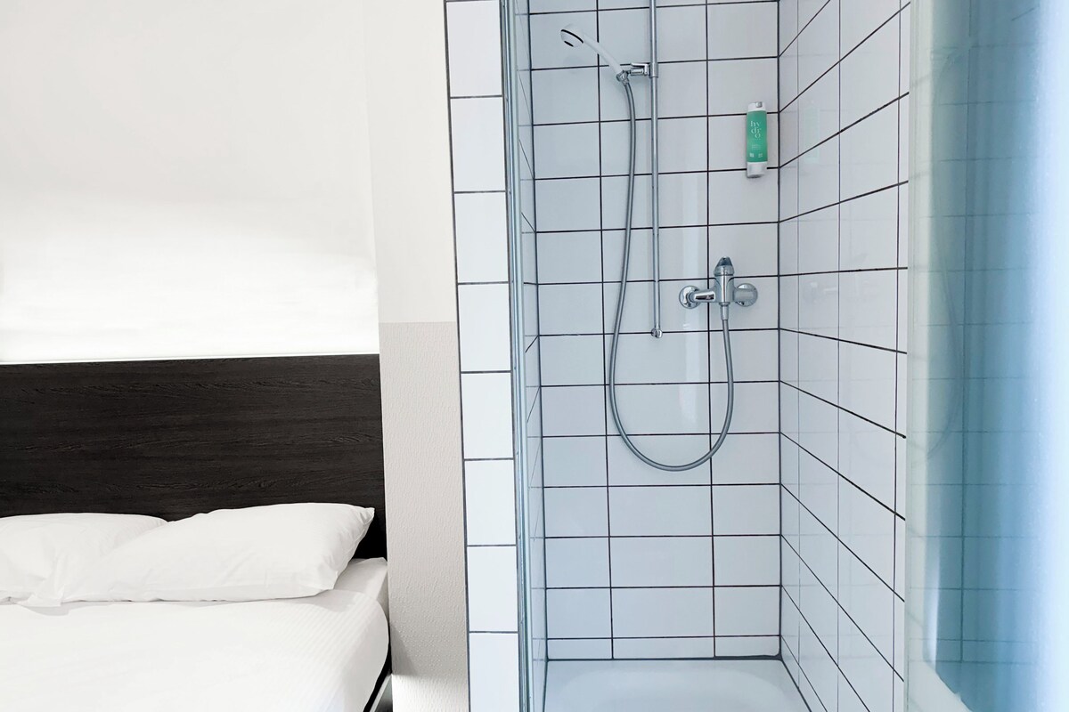 Een moderne badkamer met een douchecabine van glas, witte betegelde wanden en een bed met witte beddengoed.
