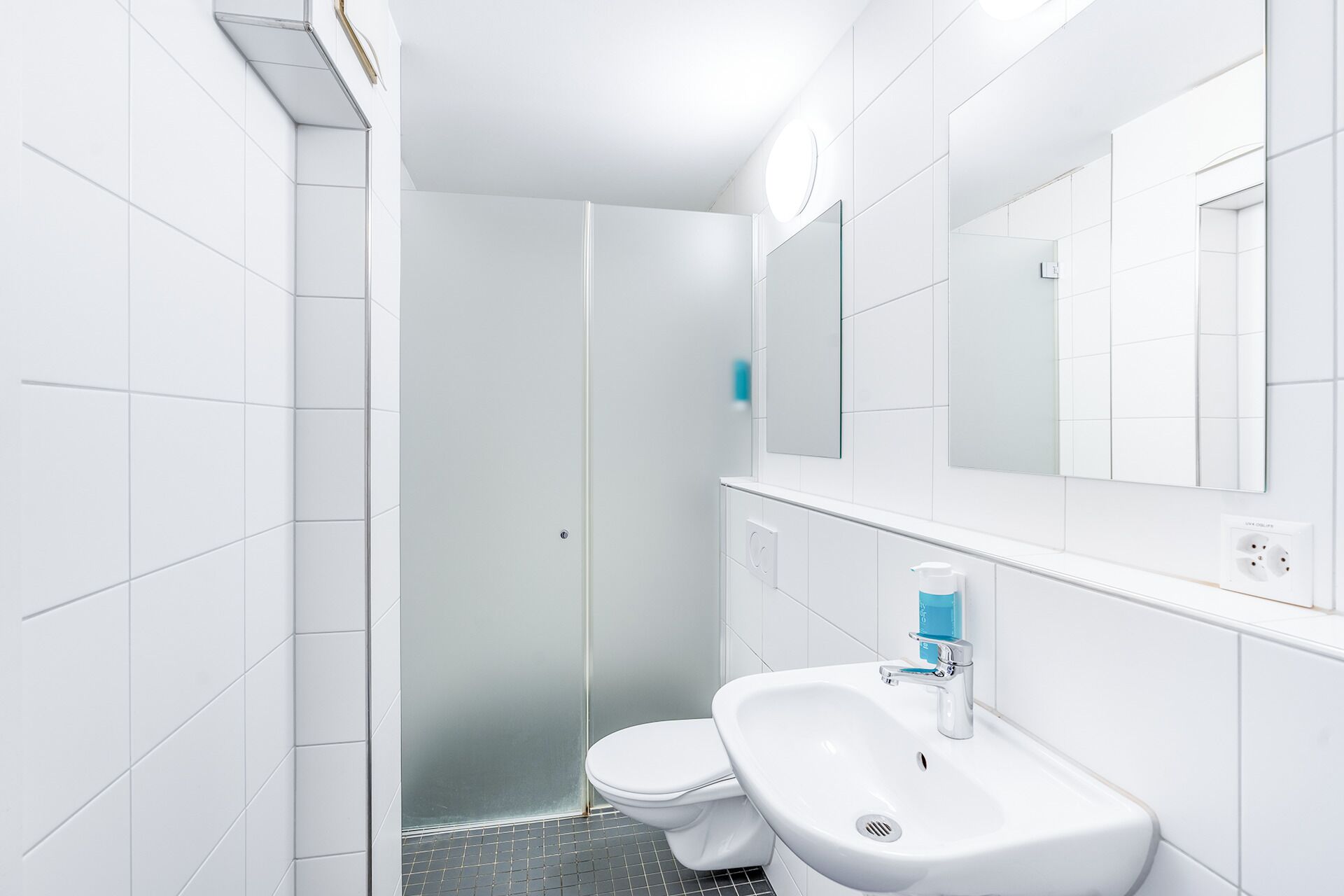 A modern bathroom with white tiles, a glass shower enclosure, a white toilet, and a sink with a blue soap dispenser.