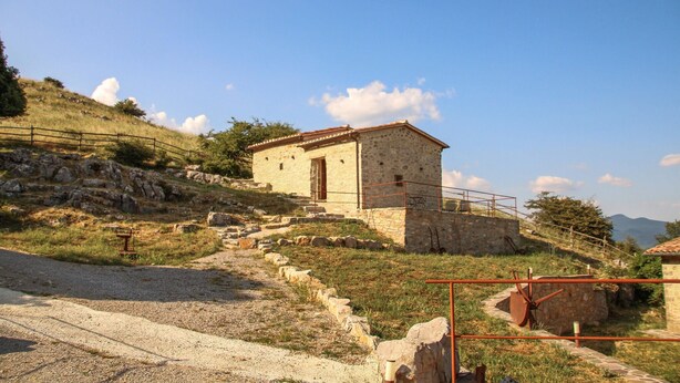 Un edificio in pietra con tetto in tegole rosse, posto su una collina con un sentiero sterrato che vi conduce.