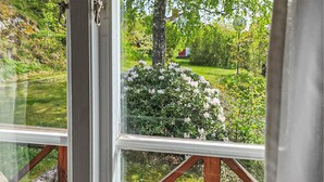 A view through a window with a wooden railing, a lush garden with various plants and flowers, and a clear sky. - Lovely home in Gränna with WiFi (Gränna)