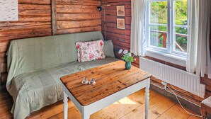 A cozy cabin room with a wooden floor, a green sofa with floral cushions, a wooden table with a vase of flowers, and a window with a view of greenery. - Lovely home in Gränna with WiFi (Gränna)