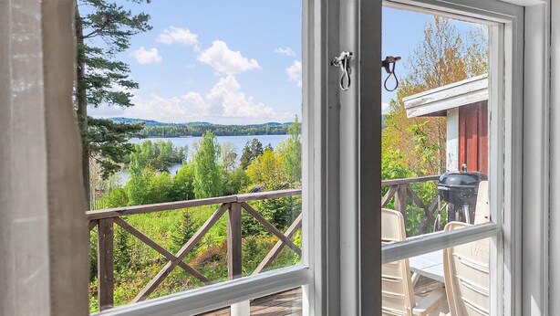 A view from a window with a wooden deck overlooking a lake, a red building, and a grill. - Lovely home in Gränna with WiFi (Gränna)