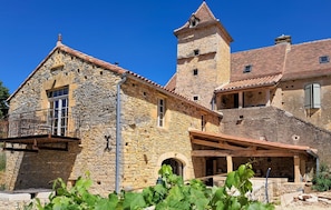 Un edificio de piedra con una torre, un balcón y un viñedo al frente.