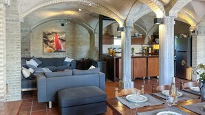 Un espacio habitable amplio con un sofá y reposapiés, una mesa de comedor lista para cuatro personas, y una cocina al fondo.
