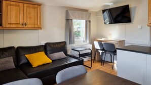 Un espace de vie compact comprenant un canapé en cuir noir, un petit bureau en bois, un téléviseur fixé au mur et une fenêtre avec des rideaux.