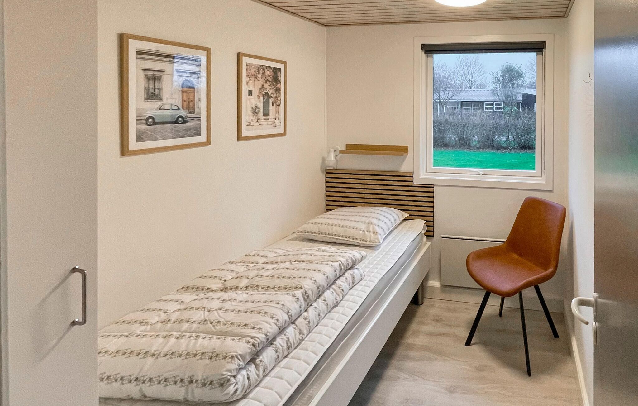 Een kleine kamer met een bed, een stoel, een raam en twee foto's in lijsten aan de muur.