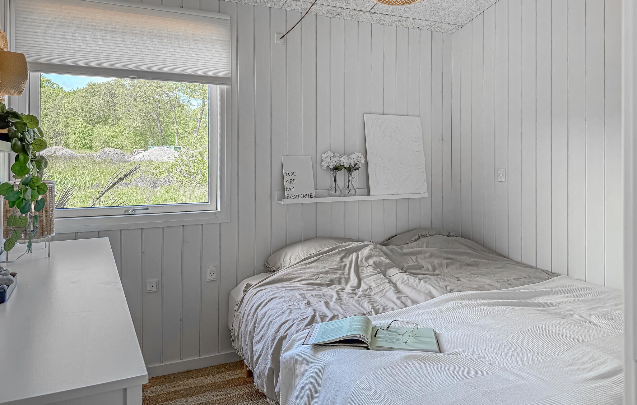 A bedroom with a bed, a bedside table, a window, and a wall-mounted shelf with decorative items.