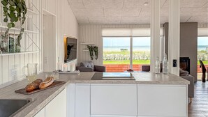 A modern kitchen with a large island, a dining area with a view of the beach, and a living room with a sofa and a TV.