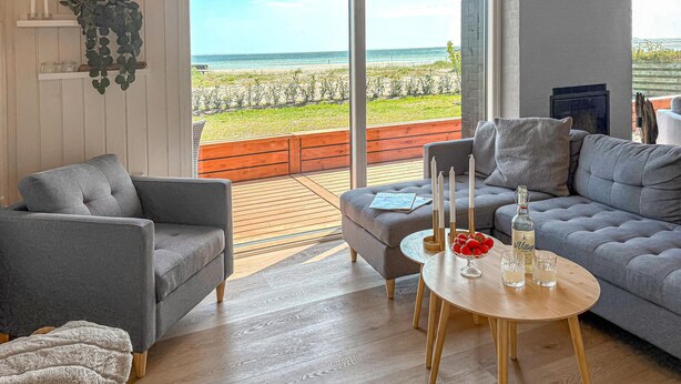 A modern living room with a grey sofa, a grey armchair, and a wooden coffee table with candles and a bottle. A large window offers a view of the beach and ocean.