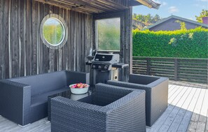 Une terrasse extérieure aménagée avec des meubles en osier, un barbecue et une vue sur une haie verdoyante et une maison en arrière-plan.