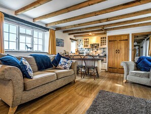 Un vaste salon comprenant un canapé et des fauteuils, des poutres en bois au plafond, un coin cuisine avec des meubles en bois et une table à manger.
