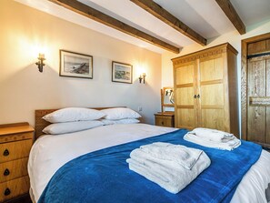 Un dormitorio con cama, colcha azul, almohadas blancas, cabecero de madera, armario de madera y dos luces fijadas a la pared.