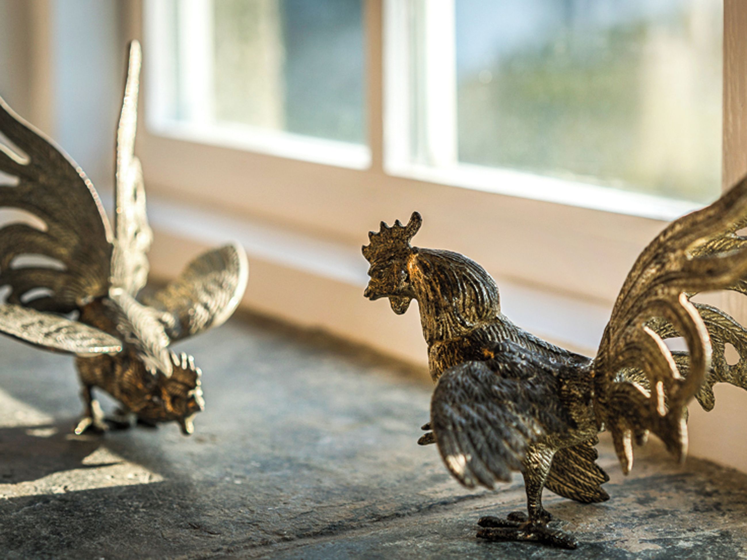 Gros plan sur des sculptures de coqs en métal ouvragé, posées sur un rebord de fenêtre.