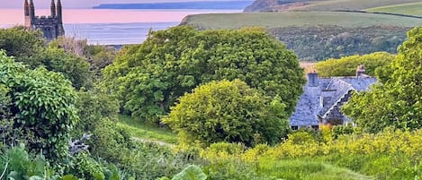 A coastal landscape with a historic tower, lush greenery, and a house with a steep roof.