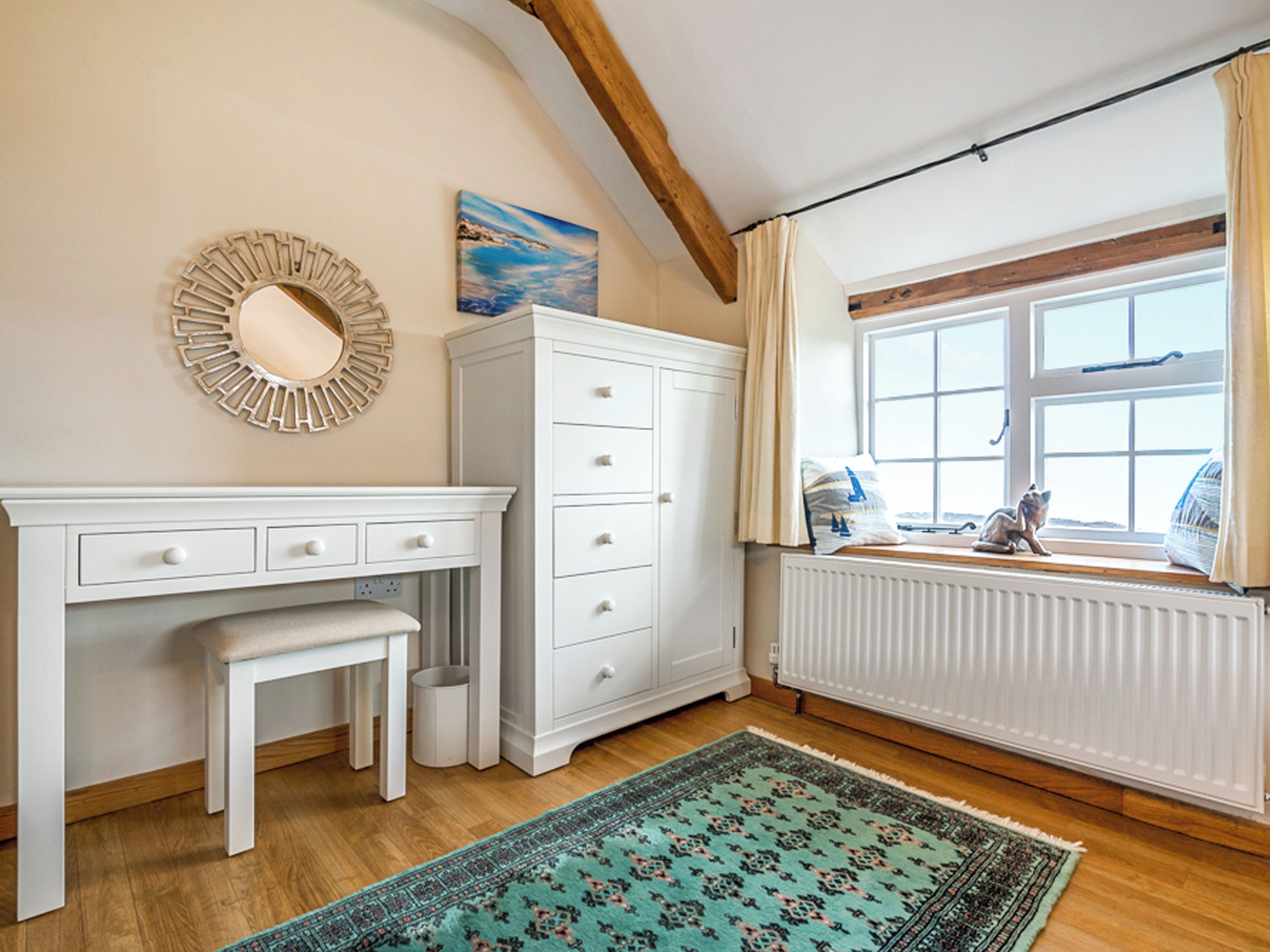 Een slaapkamer met een houten vloer, een witte ladekast, een bankje, een spiegel, een raam met gordijnen en een vloerkleed.