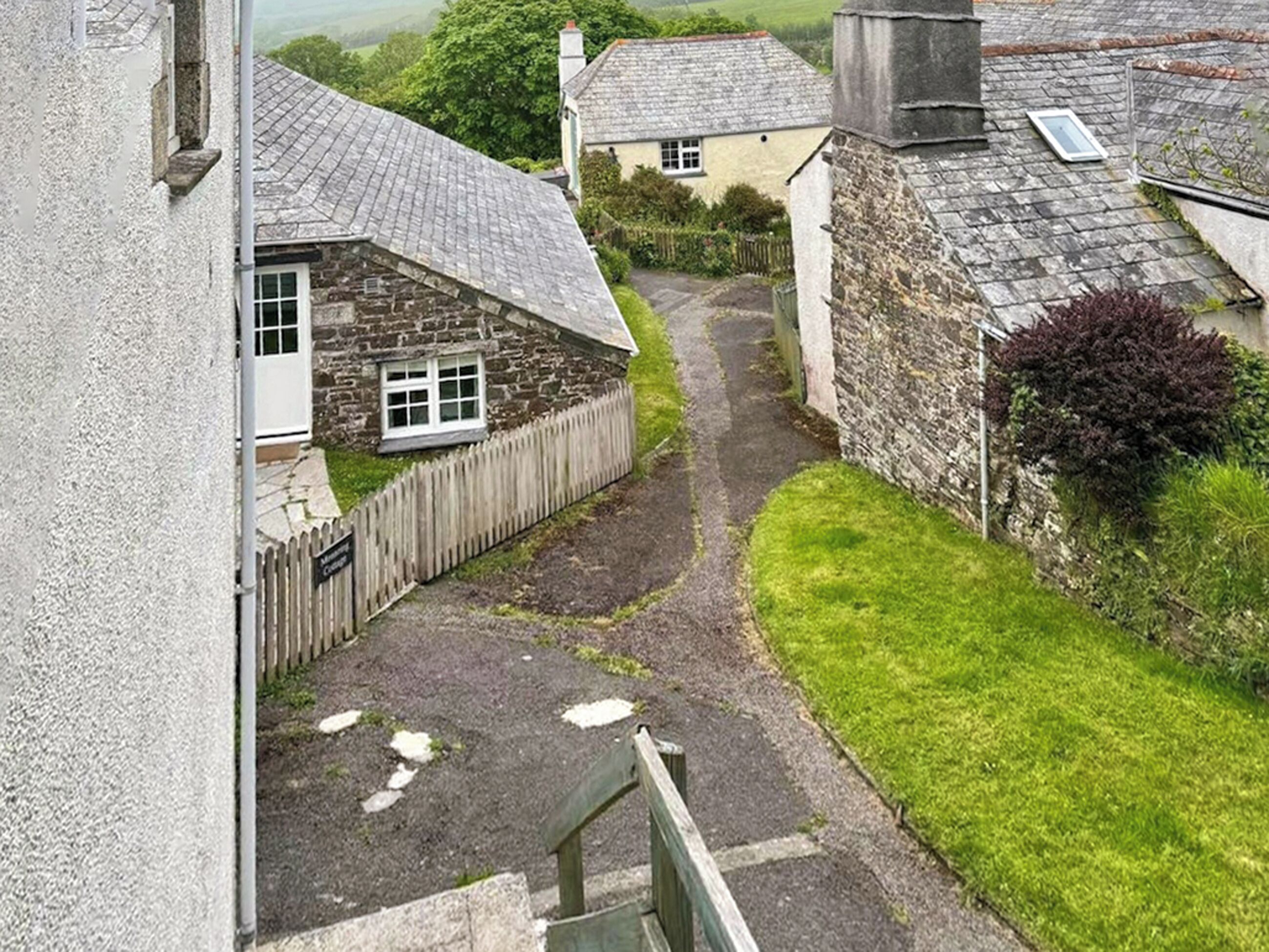 Un estrecho sendero de piedra conduce a una casa de piedra con puerta y ventana blancas, rodeada de follaje y otras construcciones de piedra.