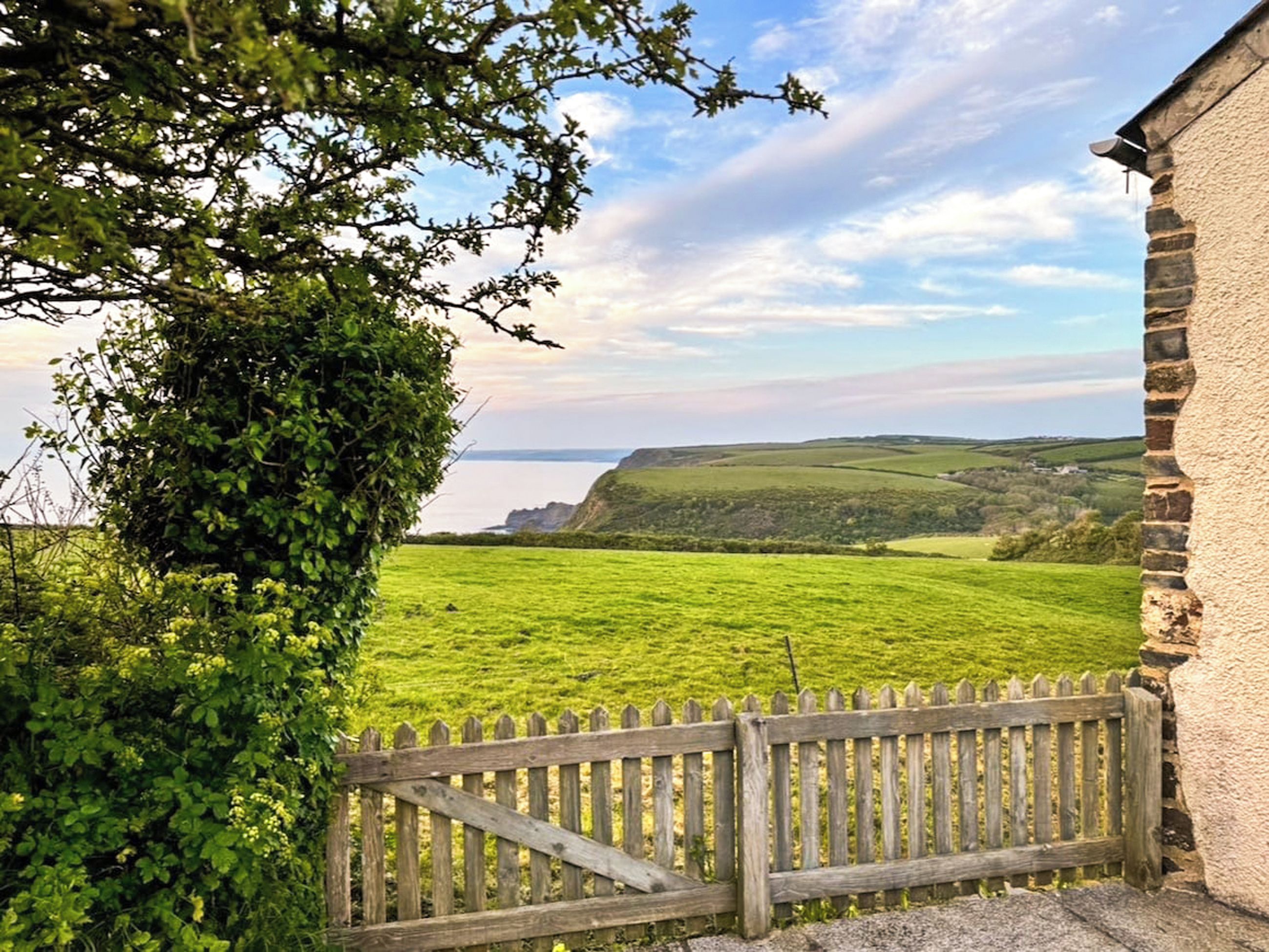 A view of a coastal landscape with a wooden gate, a stone building, and a lush green field.