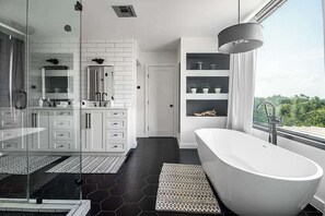 A modern bathroom with a freestanding bathtub, white cabinetry, and a large window offering a view of greenery. - Luxury 4-Bedroom Container Home (Fredericksburg)