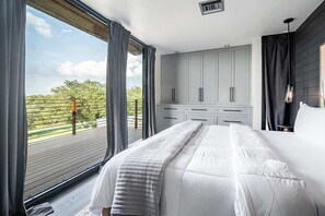 A bedroom with a large bed, a wardrobe, and a view of a deck and greenery. - Luxury 4-Bedroom Container Home (Fredericksburg)