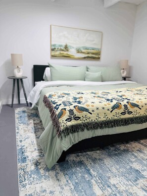 A bedroom with a bed, a nightstand, a lamp, and a painting on the wall. - Luxury 4-Bedroom Container Home (Fredericksburg)