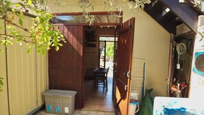 A narrow outdoor corridor with a wooden door, a small table, and a chair. A plastic container is on the ground.