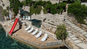 En pool av sten med en röd vattenrutschkana, solstolar och en stenmur.