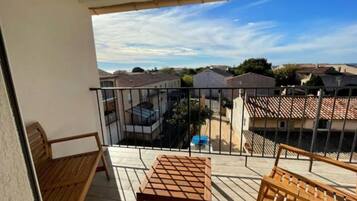 Un balcon avec une table et des chaises en bois, donnant sur un quartier résidentiel avec des maisons et un ciel dégagé.