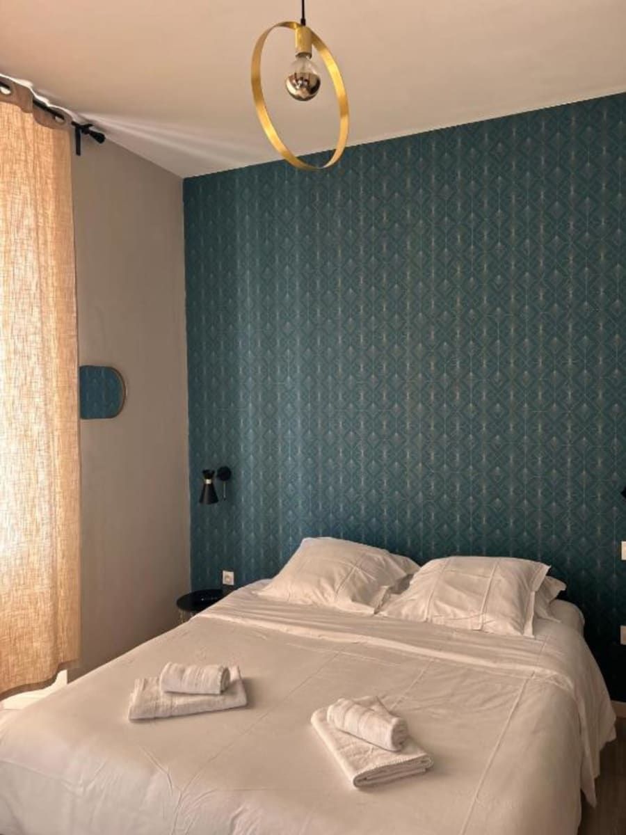 A neatly made bed with white linens and towels, a round gold light fixture, and a blue patterned wall.