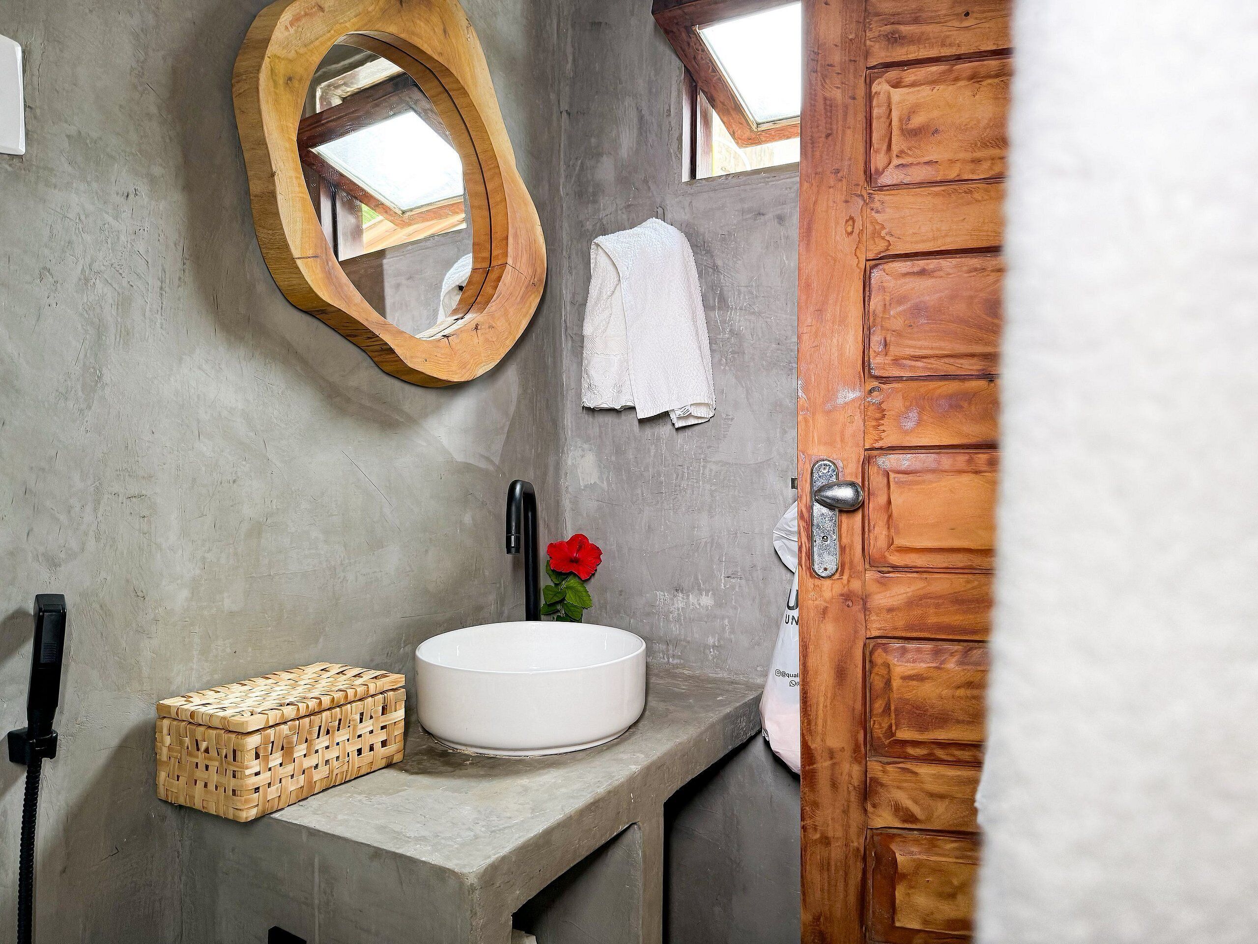 Een badkamer met een beton aanrechtblad, een spiegel met houten lijst, een witte wasbak, een rode bloem en een houten deur.