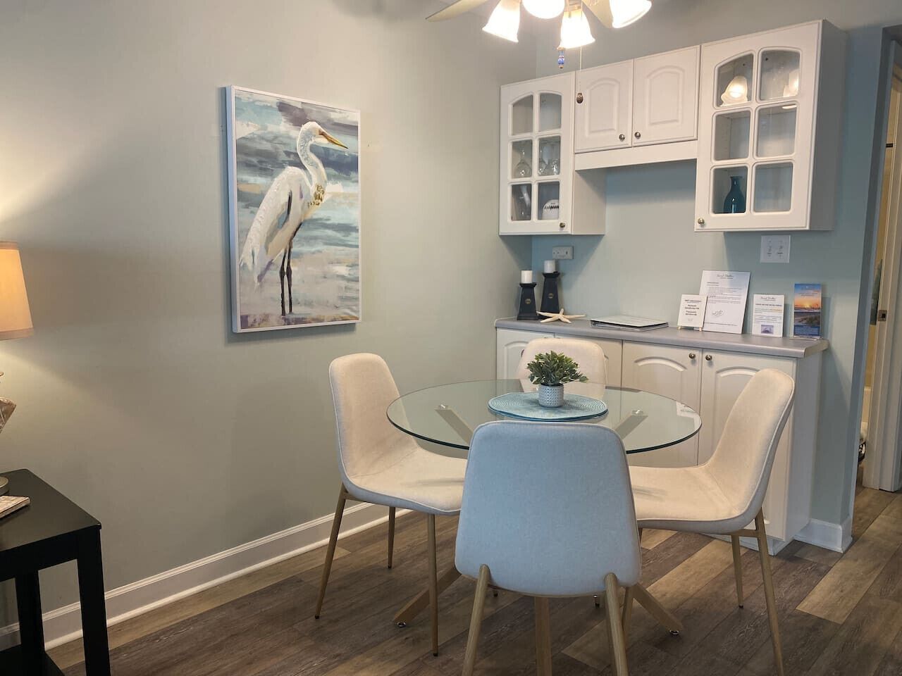 A dining area with a glass table, two chairs, a painting of a bird, and white cabinets with glass doors.