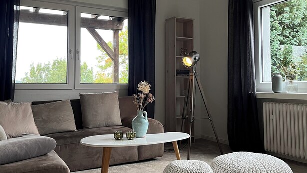 A modern living room with a sofa, a coffee table, and two ottomans. There is a floor lamp and a bookshelf in the room. - Natural paradise at the gateway to the Ruhr area: Idyllic temporary home on the Heierberg (Hattingen)